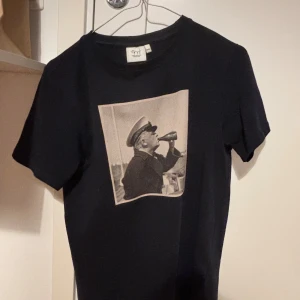 Svart t-shirt från Emmamaiina - Svart t-shirt från Emmamaiina med ett stort tryck framtill som föreställer en person i uniform som dricker ur en flaska. Klassisk rund halsringning och korta ärmar. Perfekt för dig som gillar unika motiv och stilrena plagg.