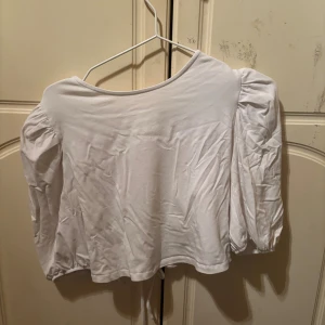 Vit blus med puffärmar och snörning - Säljer en vit blus med puffiga långärmar och snygg snörning i ryggen. Toppen har en rundad halsringning och är croppad i modellen. Materialet är mjukt och känns som bomull, perfekt för dig som gillar trendiga detaljer och en feminin look.