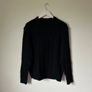 Mörkblå stickad tröja från Uniqlo - Mörkblå stickad tröja från Uniqlo med grovt kabelmönster och rund hals. Tröjan har långärmade ribbade muddar och är tillverkad av en blandning av akryl, polyester, polyamid och ull. Perfekt för dig som gillar klassisk stil med lite edge. 4E10