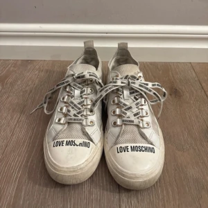 Love Moschino vita sneakers med snörning - Snygga vita sneakers från Love Moschino med tydlig logga på tån och på snörena. Skorna har mesh-paneler, guldfärgade öljetter och platt sula. Perfekt för dig som gillar streetstyle och vill sticka ut med coola detaljer.
