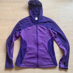 Lila fleecejacka med huva från Patagonia - Snygg lila fleecejacka från Patagonia med huva och hel dragkedja framtill. Jackan har två sidofickor med dragkedja och broderad logga på framsidan. Tillverkad i mjuk polyester med inslag av elastan för extra komfort. Perfekt för chill eller utomhusaktiviteter.