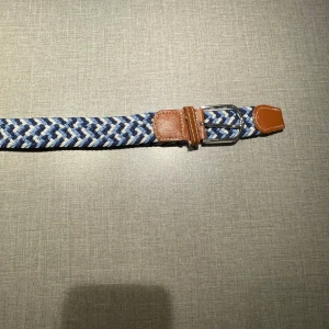 Blått flätat bälte med skinn-detaljer - Snyggt flätat bälte i blå, vit och grå med bruna skinn-detaljer vid spännet och ändarna. Bältet har ett klassiskt silverfärgat spänne och en cool, randig look som passar till jeans eller chinos. Perfekt accessoar för att lyfta din outfit.