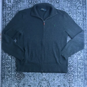 Mörkgrå kofta från Polo Ralph Lauren - Snygg mörkgrå kofta från Polo Ralph Lauren med dragkedja och ribbad krage. Tröjan har långärmade ärmar och ett grönt broderat Polo-logo på bröstet. Passar mer som L.