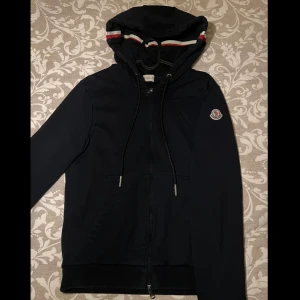 Marinblå Moncler hoodie med huva - Svart hoodie från Moncler med dragkedja, huva och snörning. Klassisk Moncler-logga på ärmen och snygga rödvita ränder på huvan. Mjuk bomullskvalitet och praktiska fickor framtill. Perfekt för dig som gillar stilrena och exklusiva streetwear-plagg.