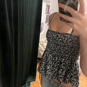Svart blommig linnetopp med volang - Säljer en svart linnetopp med vita blommor och smockad överdel. Toppen har tunna axelband och en volangig nederdel som ger en söt och avslappnad vibe. Perfekt för varma dagar och enkel att matcha med jeans eller kjol.