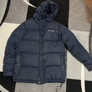 Mörkblå pufferjacka från Cross Sportswear passar från S till L stl - Säljer en mörkblå pufferjacka från Cross Sportswear med huva och snörning. Jackan har dragkedja framtill, två sidofickor med dragkedja och ett stilrent tryck på bröstet. Perfekt för kalla dagar och har en schysst oversized vibe.