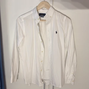 skjorta Ralph Lauren - Vit skjorta från Ralph Lauren i slim fit-modell med klassisk krage och knappar framtill. Skjortan har lång ärm och ikonisk broderad logga på bröstet. Tillverkad i mjuk bomull som känns skön mot huden. Perfekt för dig som gillar stilrena och tidlösa plagg.