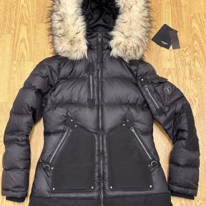 Svart dunjacka med fuskpäls från Rockandblue, modell Valencia. - Snygg svart dunjacka från Rockandblue med stor huva kantad av ljus fuskpäls. Jackan har flera dragkedjor, stora fickor framtill och en patch på ärmen. Perfekt för kalla dagar med sin vadderade design och coola detaljer. Nyskick, kvitto finns, använd ca 3 gånger, nypris 4999kr.