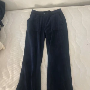 Mörkblå velourbyxor Juicy Couture XS - Mörkblå mjukisbyxor från Juicy Couture i velour med bred resårmidja och snörning. Byxorna har stora fickor fram och bak med lock och guldiga knappar. Klassisk loose passform och riktigt sköna att chilla i.