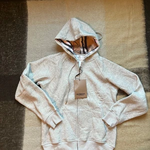 2st Burberry hoodie. Packet pris! - Burberry hoodie i både ljusgrått och svart med klassiskt rutigt foder i huvan. Tröjan har dragkedja framtill, snörning i huvan och broderad Burberry-logga på bröstet. Tillverkad i mjuk bomull för maximal komfort och stilren look.