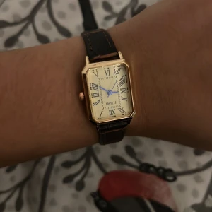 Guldig fyrkantig klocka från JUSMI - Stilren klocka från JUSMI med fyrkantigt guldigt boett, vit urtavla med romerska siffror och blå visare. Mörkbrunt läderband med synliga sömmar ger en klassisk vibe. Perfekt accessoar för dig som gillar retro och elegant design.