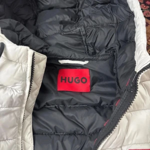 Vit pufferjacka från HUGO - Snygg vit pufferjacka från HUGO med svart foder och röda detaljer. Jackan har huva, dragkedja framtill och en röd HUGO-logga på bröstet. Insidan har en praktisk innerficka med röd dragkedja. Perfekt för kalla dagar när du vill hålla stilen.