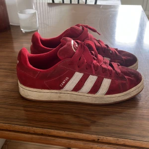 Röda Adidas Campus sneakers mocka - Säljer ett par röda Adidas Campus sneakers i mocka med klassiska vita ränder på sidorna och vit logga bak. Skorna har snörning och platt sula, samt rund tå. Perfekta för dig som gillar retrostil och vill sticka ut med färg.