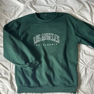 Mörkgrön sweatshirt Los Angeles - Mörkgrön sweatshirt med tryck 'Los Angeles California' i vitt framtill. Klassisk rund halsringning och ribbade muddar vid ärmslut och nederkant. Perfekt för en chill och avslappnad stil. Lite nopprig annars bra skick!