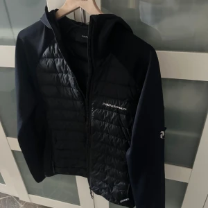  Peak Performance  - Svart pufferjacka från Peak Performance med quiltad framsida och broderad logga på bröstet. Jackan har huva, dragkedja och släta ärmar med logotyp på ena ärmen. Perfekt för kyliga dagar och snygg till streetwear passar också m