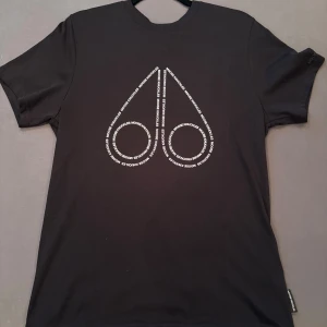 Svart Moose Knuckles t-shirt - Svart t-shirt från Moose Knuckles med stort vitt logotryck på bröstet där märkesnamnet repeteras i mönstret samt logga på armen . Klassisk passform med rund hals och korta ärmar. Äkta och allt, vill du ha mer bilder osv äre ba komma Pm. Helt oanvänd pga av att storleken ej har passat.  Passar både S och M