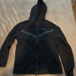 Svart Nike hoodie med dragkedja - Svart hoodie från Nike med hel dragkedja och huva. Snygg detalj med diskret V-formad rand över bröstet. Perfekt för en sportig look eller chill vardagsstil. Klassisk Nike-logga på bröstet och mjukt material.