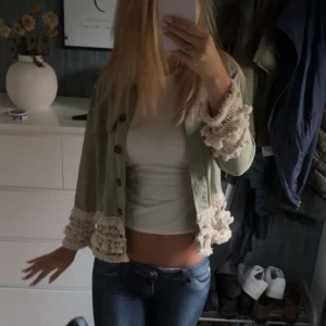 Grön overshirt med spetsdetaljer - Snygg ljusgrön overshirt med croppad passform och unika spets- och volangdetaljer vid ärmslut och nederkant. Jackan har knappar framtill och är perfekt för dig som vill sticka ut med en cool och bohemisk stil.