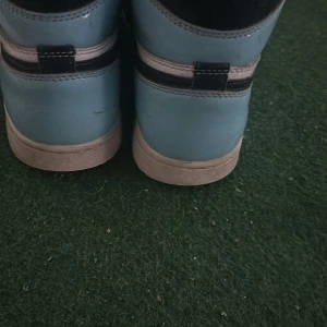 Nike Air Jordan 1 High blå/vit - Säljer ett par Nike Air Jordan 1 High i blått, vitt och svart. Skorna har klassisk Jordan-design med svart swoosh, blå detaljer och vit mittpanel. Ovandelen är i skinn och de har svarta snören samt perforerad tå. Perfekta för dig som gillar streetwear och sneakers med ikonisk stil.