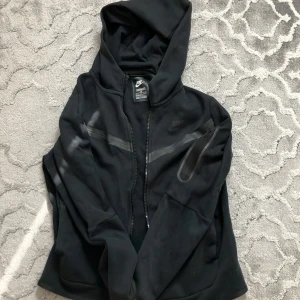 Svart Nike tech  - Svart Nike tech med hel dragkedja framtill och huva. Tröjan har en diskret logga på bröstet och en snygg detalj med en diagonal reflexrand samt en ficka med dragkedja på ärmen. Perfekt för träning eller chill.