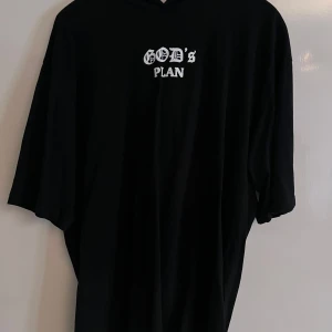 Svart oversized hoodie med tryck - Svart oversized hoodie med vit text 'God's Plan' på bröstet och stor grafisk print på ryggen med motiv av en kvinna och coolt mönster runt. Hoodien har kortare ärmar och är tillverkad i mjuk bomull. Perfekt för dig som gillar streetwear vibes.