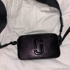 Svart axelväska från Marc Jacobs - Snygg svart axelväska från Marc Jacobs med ett stort logotypmärke på framsidan. Väskan har en justerbar axelrem . Perfekt för att bära dina nödvändigheter med stil.