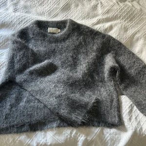 Grå fluffig stickad tröja - Mysig grå stickad tröja från Wakakuu med rund hals. Perfekt för kyliga dagar och enkel att matcha med jeans eller kjol. Långärmad och i en klassisk, rak passform.