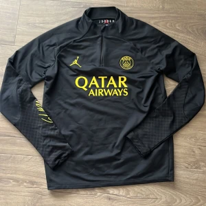 PSG långärmad träningströja Jordan XL - Svart långärmad fotbollströja från PSG x Jordan med gul logga, klubbmärke och Qatar Airways-tryck. Tröjan har halv dragkedja framtill och är i slim fit-modell. Materialet är lätt och stretchigt, perfekt för träning eller match.