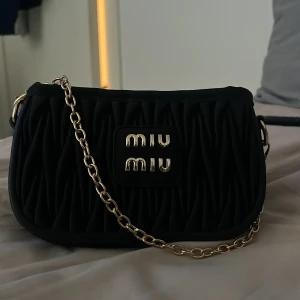 Svart axelväska från Miu Miu med kedja - Säljer en svart axelväska från Miu Miu med snyggt quiltad design och guldfärgad kedja. Väskan har ett stort Miu Miu-logo framtill och stängs med dragkedja. Perfekt storlek för det viktigaste och ger en lyxig känsla till din outfit.