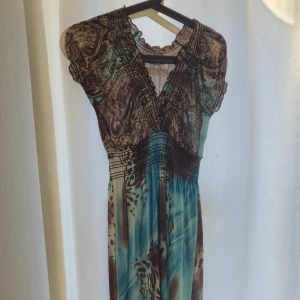 Blåbrun långklänning med leopardmönster - Säljer en långklänning i blått och brunt med leopardmönster och paisley-detaljer. Klänningen har v-ringning, korta ärmar och smockad midja som ger en snygg siluett. Materialet är tunt och flowy, perfekt för varma dagar. One size