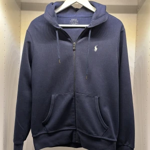 Marinblå hoodie från Polo Ralph Lauren - Snygg marinblå hoodie från Polo Ralph Lauren med dragkedja, huva och snörning. Klassisk broderad logga på bröstet och kängurufickor framtill. Tillverkad i mjuk bomullsmix, perfekt för chill dagar eller när du vill ha en clean look.