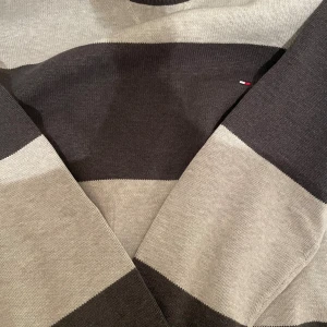 Randig tröja från Tommy Hilfiger - Snygg stickad tröja från Tommy Hilfiger i breda grå och mörkgrå ränder. Klassisk rund halsringning och diskret logga på bröstet. Perfekt för dig som gillar stilrena och tidlösa plagg.