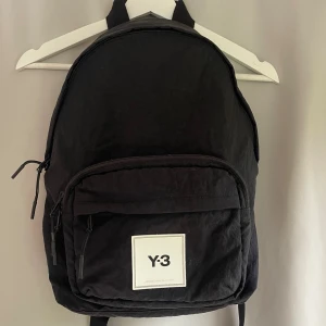 Svart ryggsäck Y-3 Adidas  - Svart ryggsäck från Y-3 Adidas Yohji Yamamoto med stilren design. Rymligt huvudfack, stor ytterficka med dragkedja och justerbara, vadderade axelremmar. Vit logotypplatta framtill ger en clean och modern look. Perfekt för skola eller cityhäng. Knappt använd, nypris 1199. 