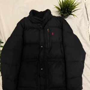 Svart pufferjacka från Ralph Lauren - Svart pufferjacka från Ralph Lauren med röd broderad logga på bröstet. Jackan har hög krage, dragkedja och knappar framtill samt två stora fickor. Insidan av kragen är fodrad i svart och jackan har rött innerfoder. Perfekt för kalla dagar.