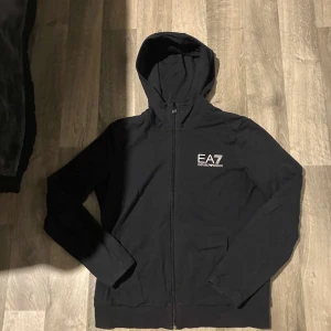 Svart hoodie från EA7 Emporio Armani -  kofta/hoodie från Armani EA7. Äkta original, köpt från auktoriserad återförsäljare. Modellen har dragkedja framtill, huva och diskret logotyp på bröstet. Passar perfekt både till vardags och träning tack vare den mjuka och bekväma bomullsblandningen. 	•	Märke: Armani EA7 	•	Storlek: L (Dam herr ochså men du behöver vara S eller M men de mer för kvinnor  	•	Färg