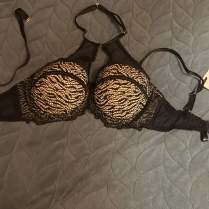 Svart spetsig korsett-topp med mesh - Säljer en svart bh från Devil med snygga spetsdetaljer och meshpartier. Bh har justerbara axelband, dekorativa ringar i metall och genomskinliga partier som ger en edgy look. Kuporna är vadderade och täckta med spetsmönster. Perfekt för dig som vill sticka ut.