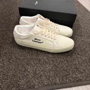 Saint Laurent beige sneakers i canvas - Snygga beige sneakers från Saint Laurent med handskriven logga på sidan. Skorna har klassisk låg profil, rund tå och platt sula. Tillverkade i canvas med detaljer i skinn och matchande beige snörning. Perfekta för en clean och stilren look.