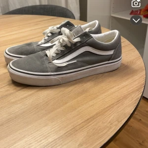 Gråa Vans Old Skool sneakers - Säljer ett par klassiska Vans Old Skool sneakers i grå mocka och canvas med vit sula och vit sidostrip. Skorna har snörning och den ikoniska gummisulan med rutmönster. Perfekta för en avslappnad streetstil.