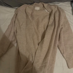 Beige kofta från H&M i ullmix - Mysig beige kofta från H&M i mjuk ullblandning. Öppen framtill utan knappar, med ribbade muddar vid ärmslut och hals. Perfekt för lager-på-lager och chill dagar. Enkel och stilren design som passar till det mesta.