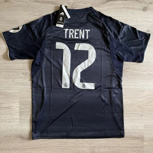 Real Madrid Trent 12 borta fotbollströja - Snygg Real Madrid fotbollströja från Adidas med Trent och nummer 12 på ryggen. Tröjan är mörkblå med tunna ränder, vita detaljer och klubbmärke på bröstet. Tillverkad i lätt funktionsmaterial med korta ärmar och klassiska Adidas-ränder på axlarna.