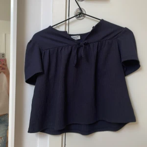 Mörkblå blus med knyt från Kappahl - Supersöt mörkblå blus från Kappahl med korta ärmar och knyt framtill vid halsen. Blusen har en lös passform och är gjord i ett lätt, strukturerat tyg som ger en skön känsla. Perfekt att styla med jeans eller kjol för en chill look.