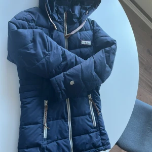 Mörkblå pufferjacka från North Bend - Snygg mörkblå pufferjacka från North Bend med huva och vita dragkedjor. Jackan har två fickor med flätade detaljer på dragkedjorna och justerbara snören i huvan. Perfekt för kalla dagar och riktigt skön att bära.