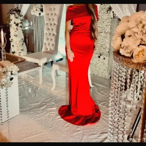 Röd axelbandslös långklänning - Säljer en elegant röd långklänning med axelbandslös design och figurnära passform. Klänningen har en snygg släp och markerad midja, perfekt för dig som vill sticka ut. Materialet har en lätt glansig finish som ger en lyxig känsla.