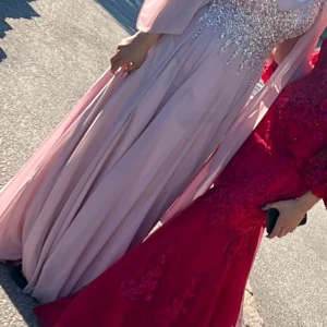 Ljusrosa långklänning med paljetter - Elegant ljusrosa långklänning med glittriga paljettdetaljer upptill och svepande kjol. Klänningen har en vacker omlottkänsla och vida ärmar som ger en drömmig look. Perfekt för bal eller festliga tillfällen.