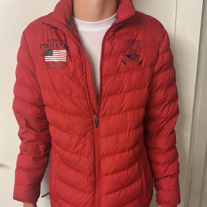 Röd pufferjacka från Vinson Polo Club - Riktigt snygg röd pufferjacka från Vinson Polo Club med broderade loggor och amerikansk flagga på bröstet. Jackan har dragkedja framtill, hög krage och quiltad design. Perfekt för kyliga dagar och ger en sportig vibe.
