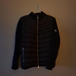 Svart pufferjacka från Lagency S - Svin snygg Svart pufferjacka från Lagency i storlek S. Jackan har quiltad framsida och rygg, stickade ärmar och krage samt dragkedja framtill och på fickorna. Klassisk och stilren look med Lagency-logga på ärmen. Det är ett litet hål vid vänstra handleden, därför priset. Det är inget man lägger märke till. Den sitter jätte bra, och passar dom som är runt 165-175cm. 