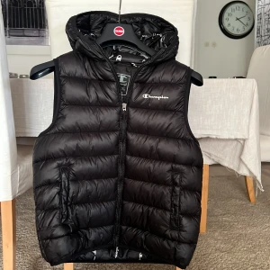 Svart dunväst från Champion XL - Svart dunväst från Champion med huva och dragkedja framtill. Västen har två fickor med dragkedja och quiltad design. Perfekt för lager-på-lager och streetstyle. Champion-logga på bröstet ger en sportig vibe. Som en XS vuxen 