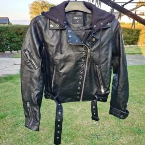 Svart skinnjacka med hoodie FB Sister - Svart skinnjacka från FB Sister i storlek S. Jackan har coola silverfärgade dragkedjor, bälte i midjan och en svart hoodie med snörning. Snygga detaljer med nitar och öglor, samt flera fickor med dragkedja. Perfekt för dig som gillar edgy streetstyle.