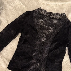 Svart spetsblus med knappar - Säljer en svart blus i genombruten spets med blommigt mönster. Djup v-ringning, långa ärmar och knappar framtill. Perfekt för dig som gillar detaljer och vill sticka ut lite extra. Snygg att styla med jeans eller kjol.