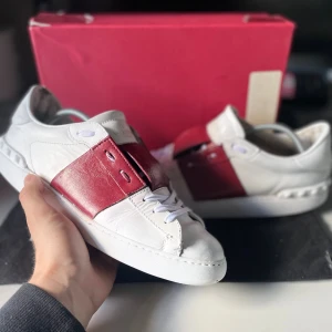 Valentino Open vit/röd - Snygga Valentino Garavani Open sneakers i vitt skinn med bred röd läderdetalj över snörningen. Klassisk rund tå, vita skosnören och ikoniska nitar baktill på sulan. Perfekta för dig som vill sticka ut med en lyxig och modern look. Acceptabelt skick, ser använda ut men har mycket kvar att ge! Skorna har inga hål eller skador endast slitna. Storlek (41~passar 42/43) box och dustbag medföljer vid köp. Nypris 7200:- mitt pris 1499:-🌟 Vid frågor är det bara att höra av sig!